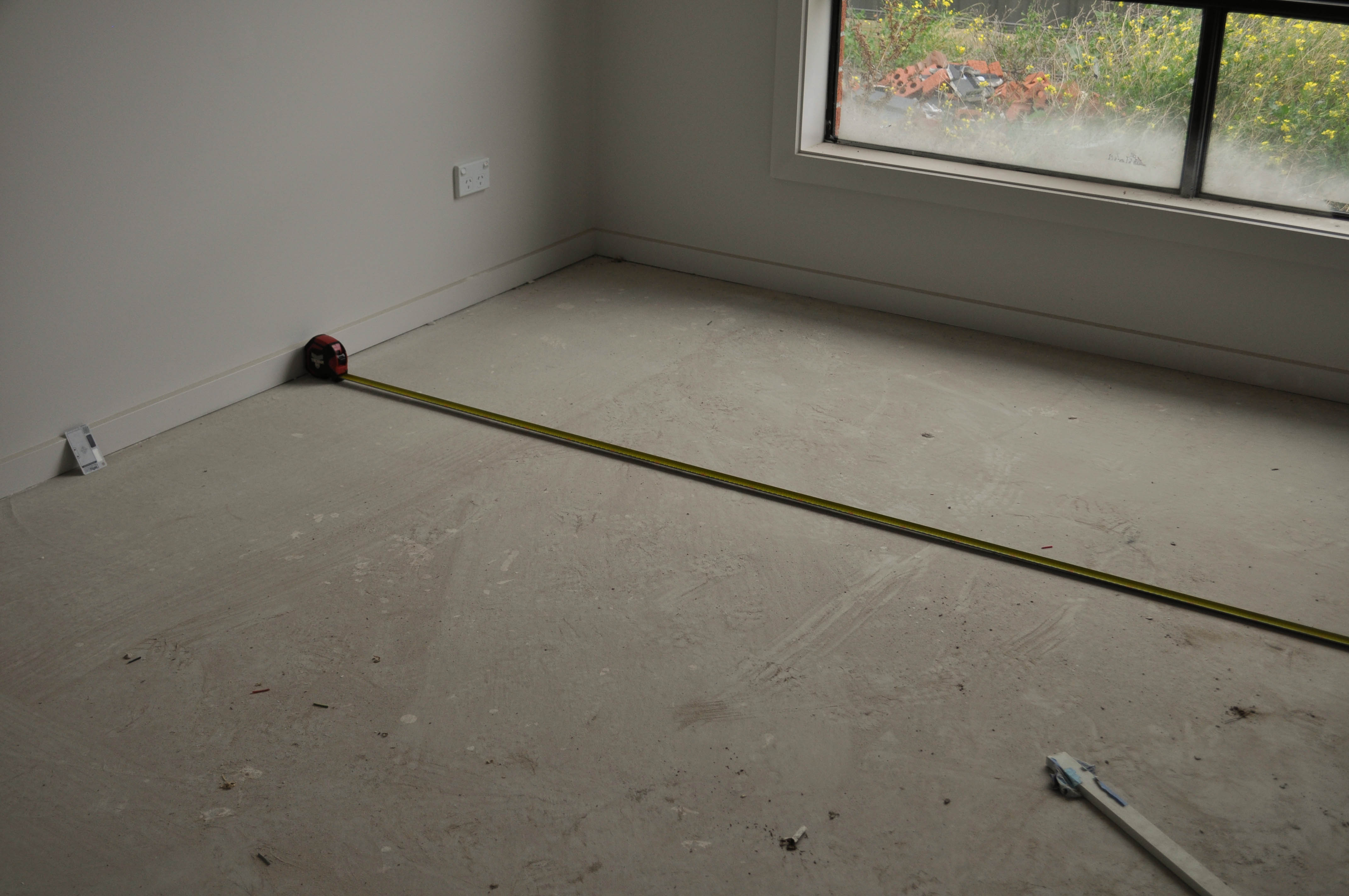 an empty room being measured for carpet quantity. A measuring tape is running across the bare concrete floor from one end of 
the room to the other end. The home is in Werribee and the measuring process was conducted by Concord Floors so that the owner could obtain a quote for the cost of carpeting
 their home if they employed Concord Floors to do the job.