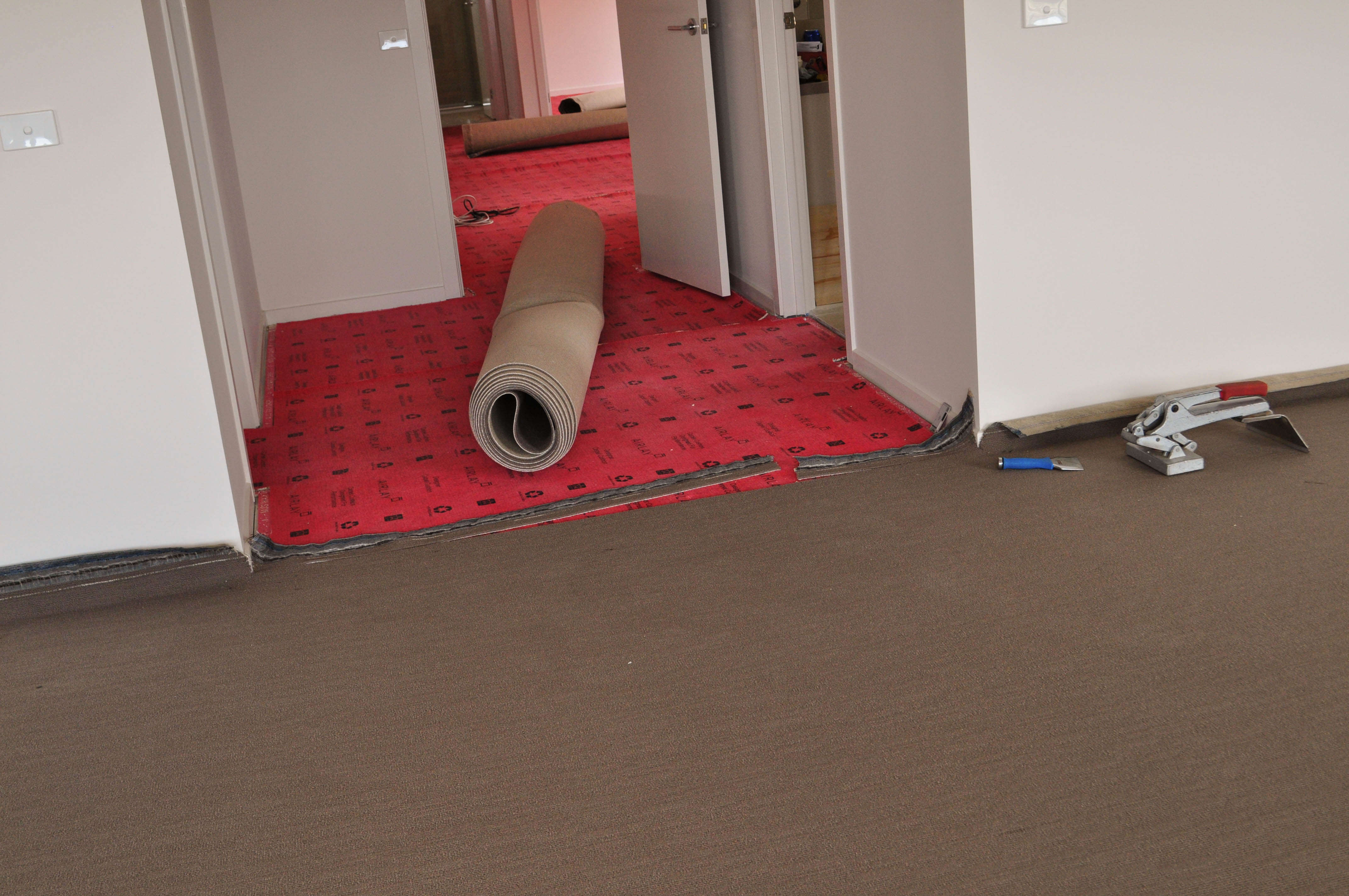 showing the work of the installer Concord Floors installing carpet in a lounge room from which a wide passage runs. The passage has gripper on the floor against the walls and underlay
 between the gripper covering the whole floor. On top of the underlay is a roll of carpet pre-cut to size ready to be laid on the floor. The lounge room has carpet stretched and tensioned ready to be joined to the passage carpet. 
 The carpet laying in full swing as part of the carpet installation procedure. The home is in Point Cook, Werribee.