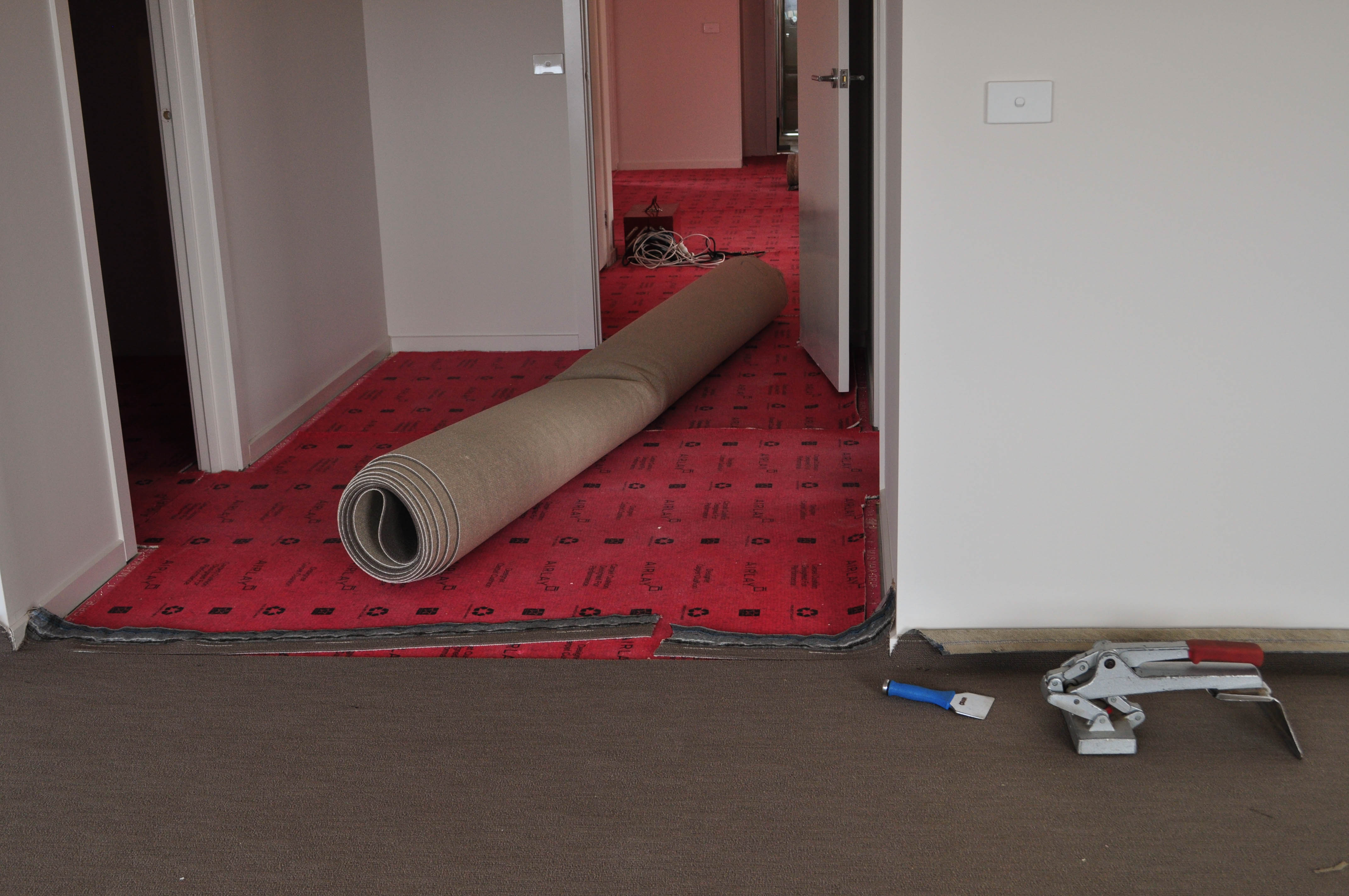showing a lounge room from which a wide passage runs. The passage has gripper on the floor against the walls and underlay between the gripper
 covering the whole floor. On top of the underlay is a roll of carpet pre-cut to size ready to be laid on the floor. The lounge room has carpet stretched and tensioned ready to be joined
 to the passage carpet. The carpet laying in full swing as part of the carpet installation procedure. The home is in Point Cook, Werribee, Vic. 3030. The carpet laying was done by 
 Concord Floors