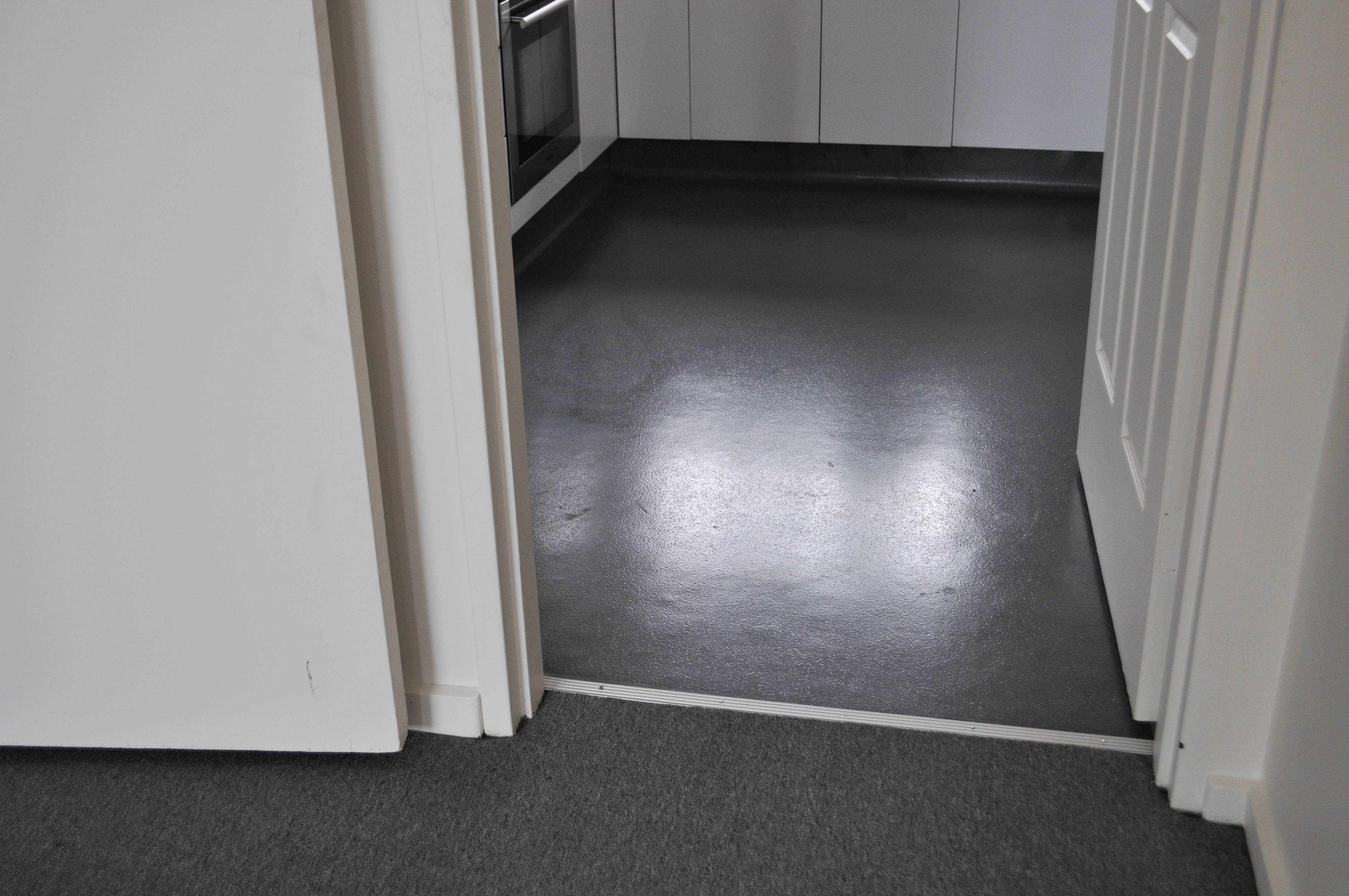 showing the carpeted loungeroom carpet of charcoal color and the kitchen with commercial, sheet vinyl flooring, type 2 metre wide, and coved 1500mm up the cabinets and walls.