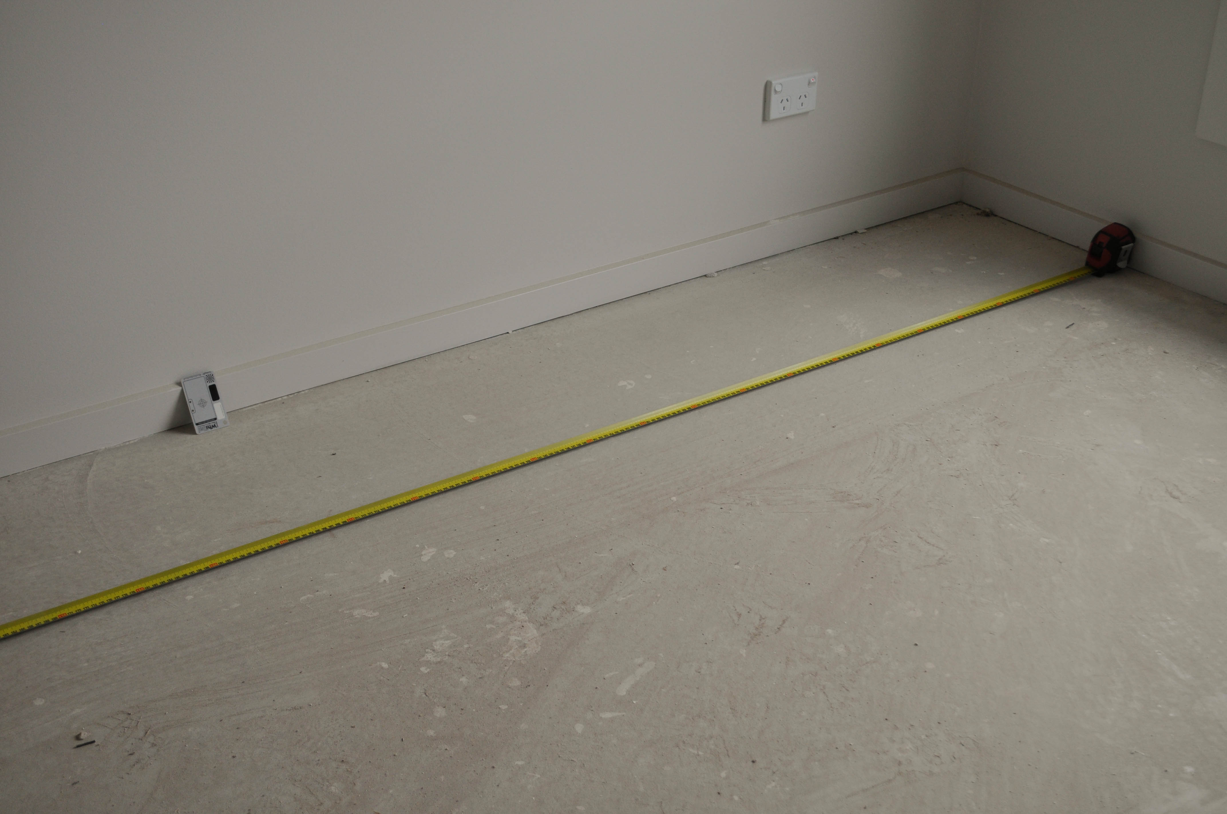 a room in a home, in Werribee, Vic 3030, being measured for the purpose of working out how much carpet is needed to cover it in a 
 carpet installation project conducted by Concord Floors. The floor of the room is bare concrete and has a measuring tape stretched over and across it. This is the initial stage of 
 the carpet installation  process.