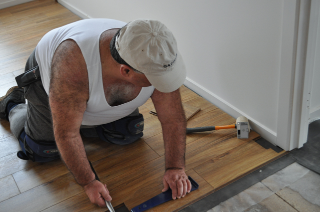 a Concord Floor's floorboard layer/installer laying 12mm thick Spotted Gum  laminate flooring, in a 
room in the suburb of Point Cook, Werribee Vic 3030. He is kneeling on the laid boards and tapping in a notched floorboard into an architrave cavity that he has made by 
 using his undercutting saw.