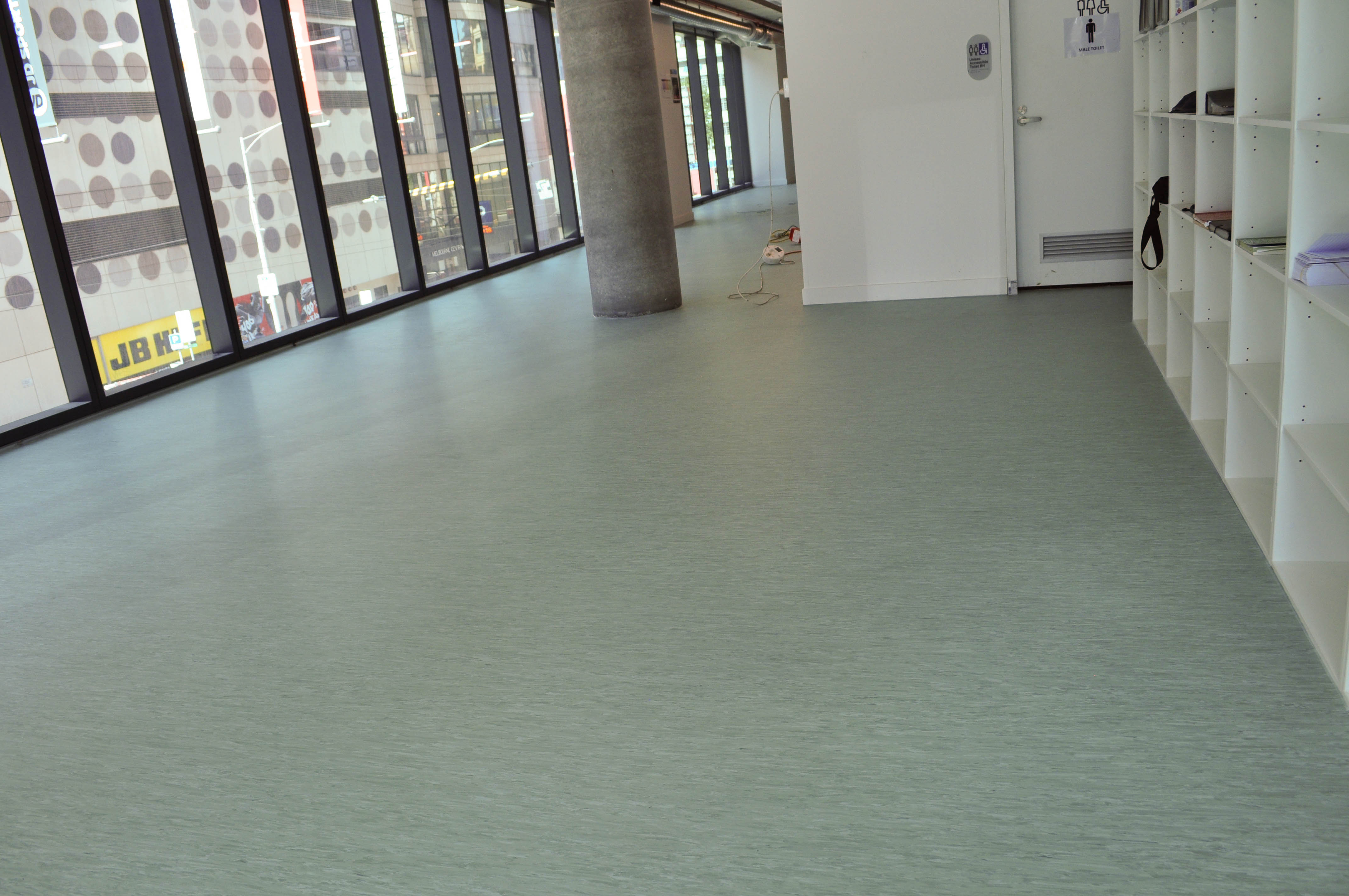 the commercial vinyl installation process completed, in an entire office block,
            in La Trobe Street, Melbourne 3000><p>The installation is by Concord Floors and is showing a very large room where the entire floor is covered by green colored commercial vinyl.
            The room has on it's left side large charcoal framed windows and on the right side white, pidgeon-holed shelving.
