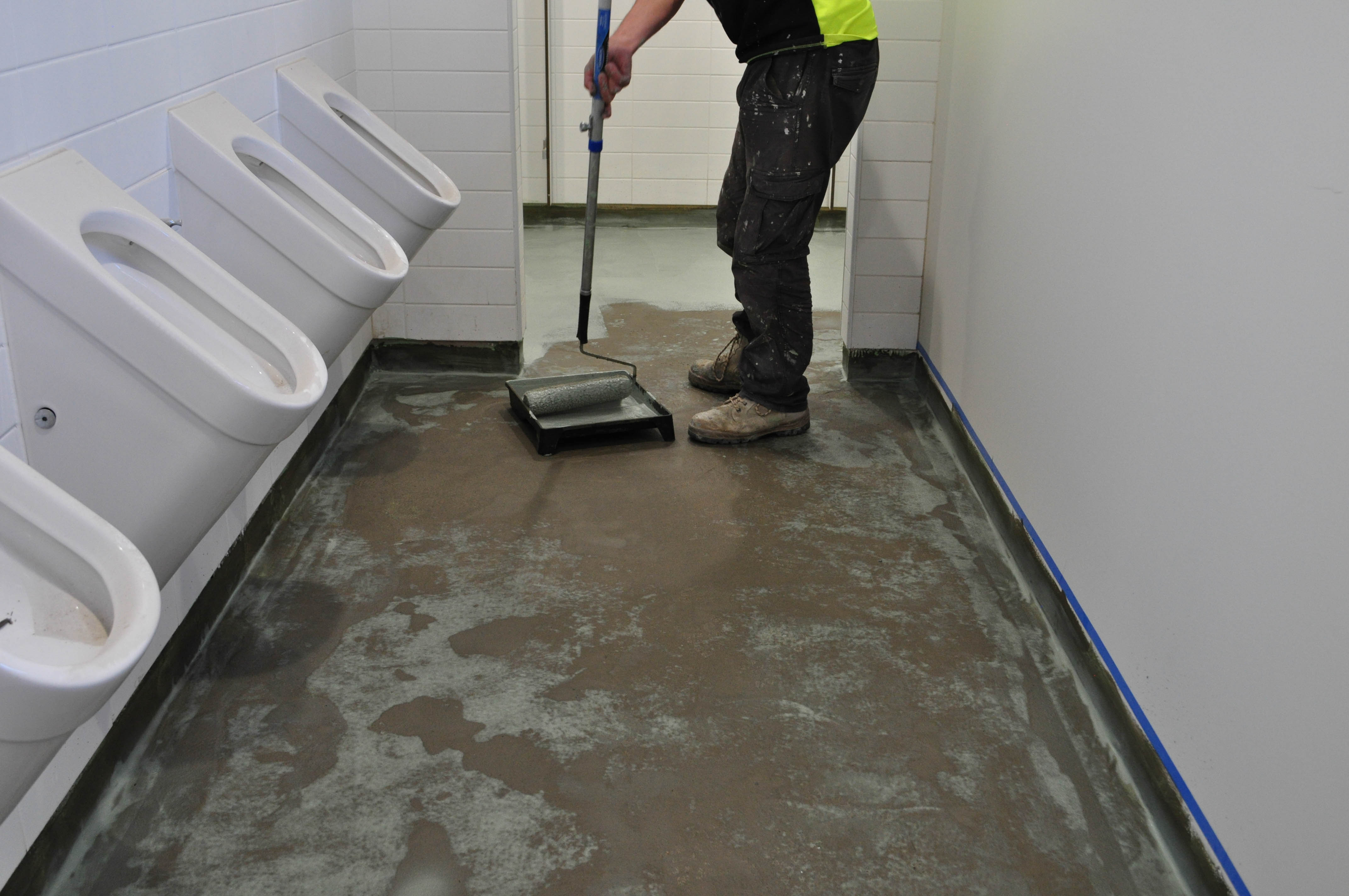 showing a toilet block floor undergoing floor preparation by Concord Floors. The room has 3 toilet pans protruding 
	  from the wall on the left hand side, the floor evenly smeared with a chemical and a vinyl layer smearing the floor with the chemical from a paint tray.