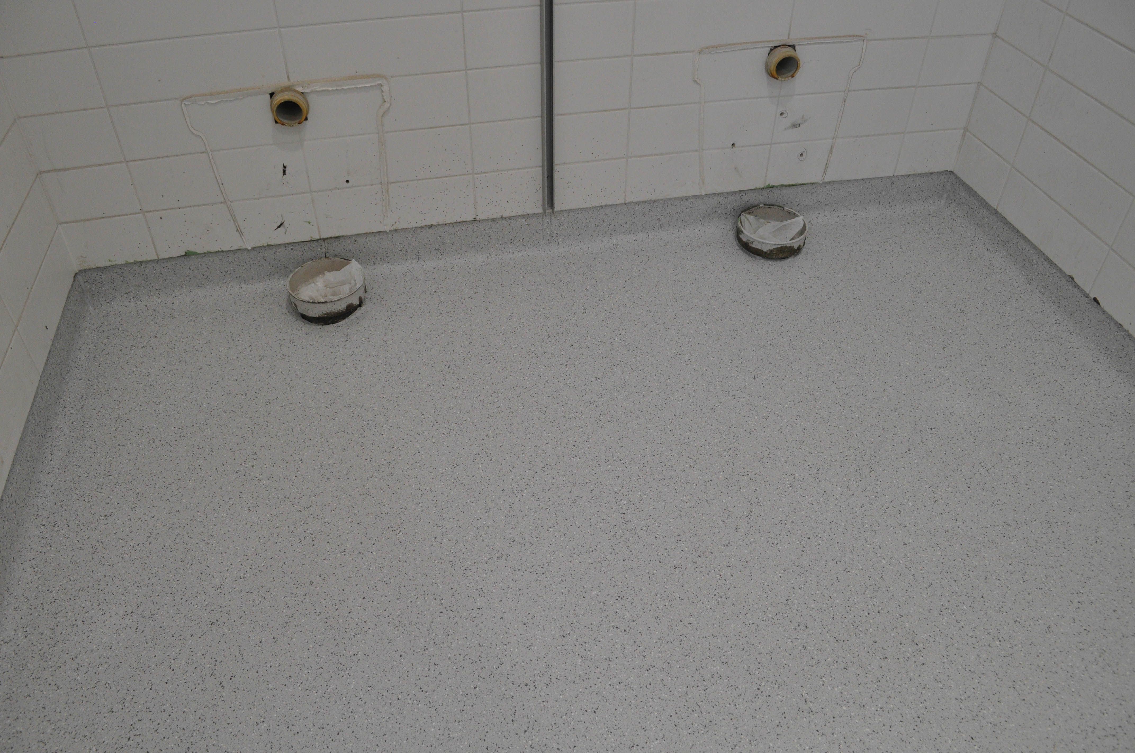 an empty room, a toilet, with exposed newly installed with vinyl coving 150mm up the wall. The vinyl surface is a light 
grey color. The walls are tiled with a white tile, that rise upwards.