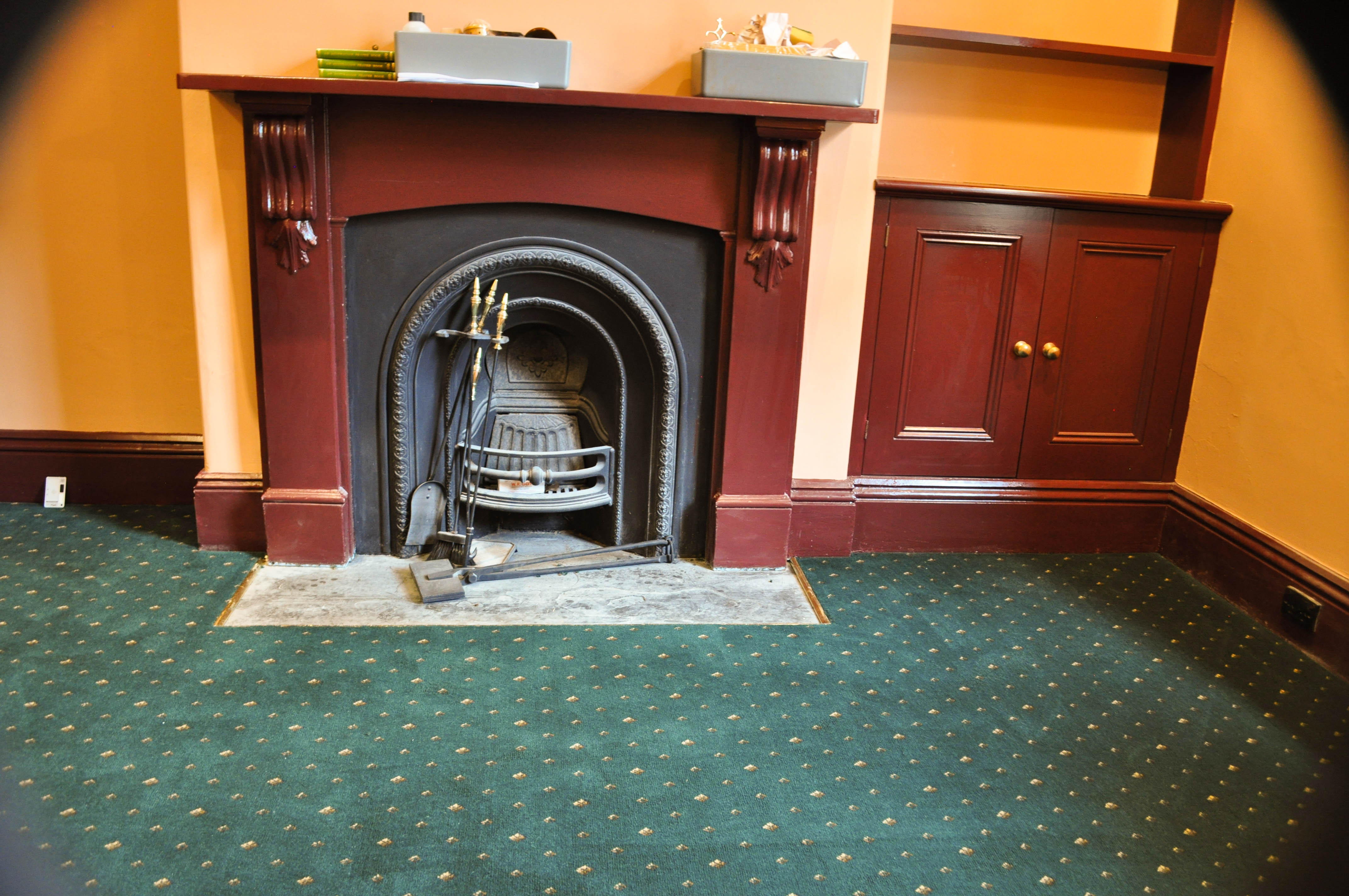 a room in the interior of a period home utilizing, a green carpet color and a period style of patterned carpet it being an aximnister carpet.
 The carpet meets a tan fireplace with a stone hearth floor. The walls are painted a deep orange color, which is period decor. The scheme is triadic, utilizing all three
 primary colors, red, blue and yellow. The home is in Melbourne, Vic 3000 and the carpet installed by Concord Floors.