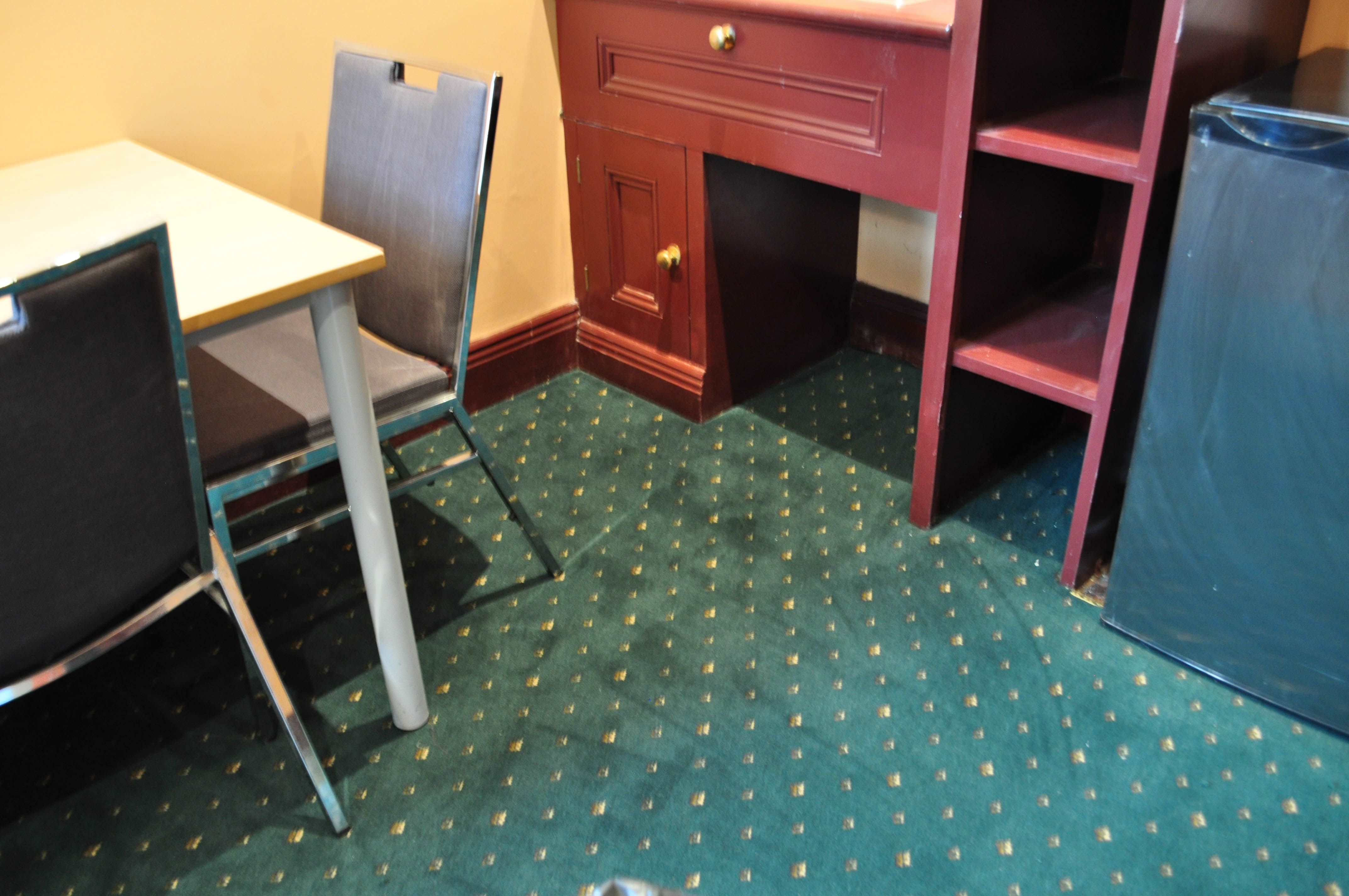 a room in the interior of a period home utilizing, a green carpet color and a period style of patterned carpet it being an aximnister carpet.
 The carpet meets a tan kitchen cabinet a simple table and chair. The walls are painted a deep orange color, which is a period decor. The home is in Melbourne, Vic 3000 and the carpet
 installed by Concord Floors.